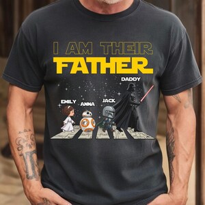 May include: A dark gray short-sleeve t-shirt featuring the yellow text I AM THEIR FATHER and DADDY. The graphic shows cartoon characters Princess Leia, BB-8, The Mandalorian, and Darth Vader walking across a crosswalk against a starry background.