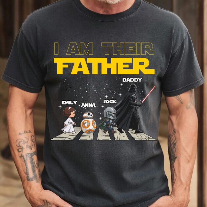 May include: A dark gray short-sleeve t-shirt featuring the yellow text I AM THEIR FATHER and DADDY. The graphic shows cartoon characters Princess Leia, BB-8, The Mandalorian, and Darth Vader walking across a crosswalk against a starry background.