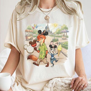 May include: Off-white t-shirt with a vibrant print of Zootopia's Nick Wilde and Judy Hopps in a theme park setting. Nick holds a Mickey Mouse-shaped snack, Judy has an ice cream cone. The background shows a castle, food stalls, and greenery.