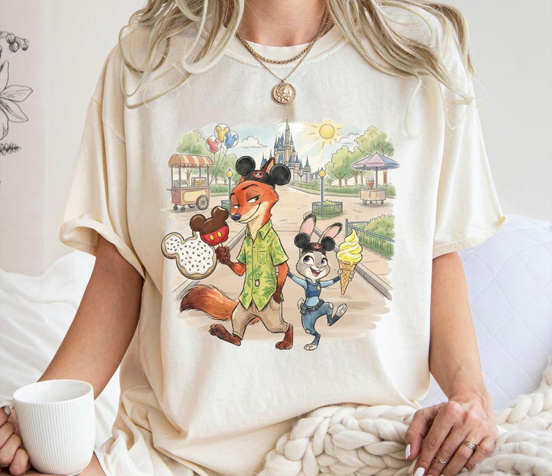 May include: Off-white t-shirt with a vibrant print of Zootopia's Nick Wilde and Judy Hopps in a theme park setting. Nick holds a Mickey Mouse-shaped snack, Judy has an ice cream cone. The background shows a castle, food stalls, and greenery.