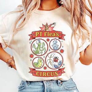 May include: Cream-colored t-shirt featuring a colorful graphic of a circus with insects. The design includes a praying mantis, ant, caterpillar, ladybug, and other insects. The text "PT Flea's Circus" is displayed in a red banner.