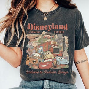 Peut inclure: T-shirt gris fonc&eacute; avec un graphisme color&eacute; des personnages de Cars de Disney et le texte "Disneyland California Est. 2012" et "Welcome to Radiator Springs". Le design comprend divers personnages de voitures anim&eacute;es et des &eacute;l&eacute;ments de paysage.