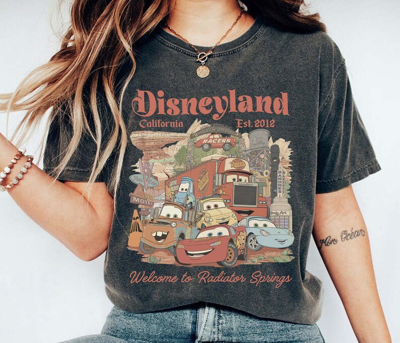 Peut inclure: T-shirt gris fonc&eacute; avec un graphisme color&eacute; des personnages de Cars de Disney et le texte "Disneyland California Est. 2012" et "Welcome to Radiator Springs". Le design comprend divers personnages de voitures anim&eacute;es et des &eacute;l&eacute;ments de paysage.