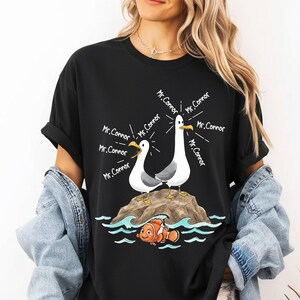 May include: Black t-shirt featuring a cartoon illustration of two seagulls on a rock, with the text "Mr. Connor" emanating from them. Below, an orange and white clownfish swims in blue waves. A fun, whimsical design.