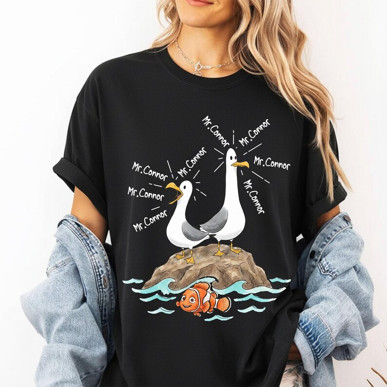 May include: Black t-shirt featuring a cartoon illustration of two seagulls on a rock, with the text "Mr. Connor" emanating from them. Below, an orange and white clownfish swims in blue waves. A fun, whimsical design.