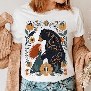 Op de afbeelding: Wit T-shirt met een gestileerde illustratie van een beer en een roodharige vrouw, omringd door bloemmotieven in goud, groen en blauw. Het ontwerp bevat een zwaard en decoratieve krullen.