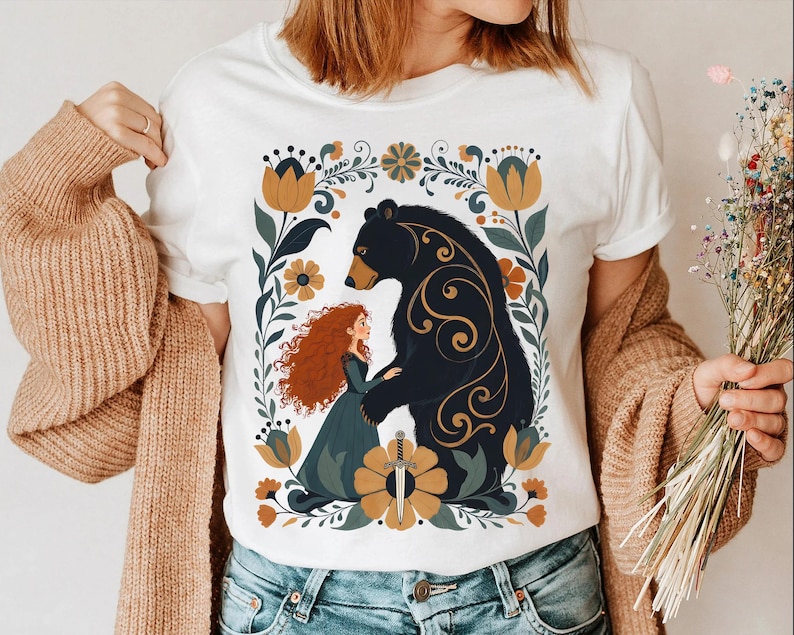 Op de afbeelding: Wit T-shirt met een gestileerde illustratie van een beer en een roodharige vrouw, omringd door bloemmotieven in goud, groen en blauw. Het ontwerp bevat een zwaard en decoratieve krullen.