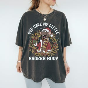 May include: Dark gray t-shirt featuring a cartoon rat wearing a Santa hat, red coat, and scarf, holding a gift. The text "GOD SAVE MY LITTLE BROKEN BODY" is printed above and below the graphic, surrounded by a wreath.