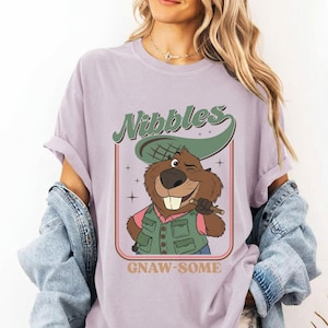 May include: A lilac-colored t-shirt featuring a cartoon beaver winking and holding a stick. The beaver wears a green vest and pink shirt. The text "Nibbles" and "Gnaw-some" are displayed in a retro font.