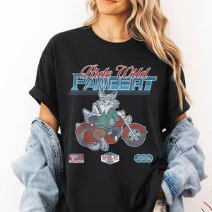Puede incluir: Camiseta negra con un conejo de dibujos animados montado en una moto roja. El texto "Ride Wild PAWBERT" est&aacute; sobre el conejo. Debajo hay tres peque&ntilde;os logotipos.