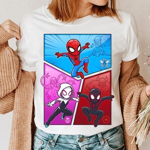 May include: White t-shirt featuring a comic book style graphic of Spider-Man and Spider-Gwen in action poses. The design includes a blue, pink, and red color scheme with additional character outlines. The shirt is worn by a person.