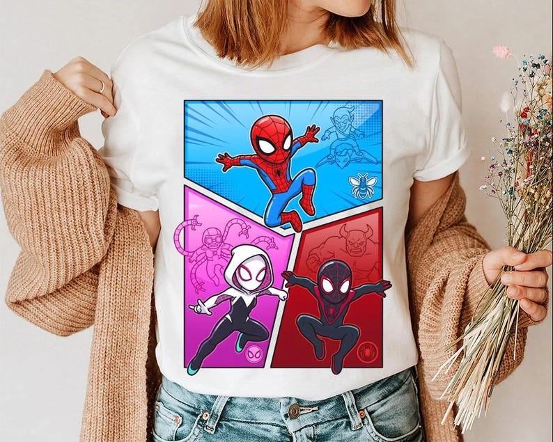May include: White t-shirt featuring a comic book style graphic of Spider-Man and Spider-Gwen in action poses. The design includes a blue, pink, and red color scheme with additional character outlines. The shirt is worn by a person.