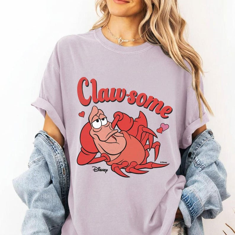 May include: A lilac-colored t-shirt featuring a cartoon image of Sebastian the crab from Disney's The Little Mermaid. The text "Claw-some" is written in red script above the crab, with small pink hearts. The Disney logo is at the bottom.