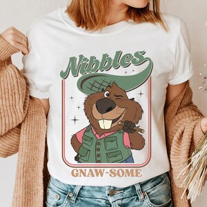 May include: White t-shirt featuring a cartoon beaver with a green hat and vest, holding a stick. The shirt has the text "Nibbles" above the beaver and "GNAW-SOME" below. The beaver is winking and smiling.