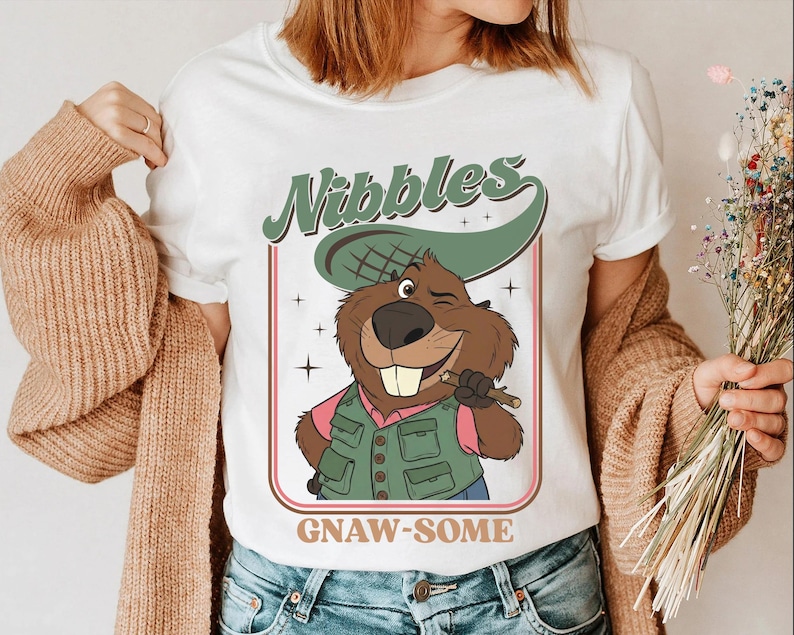 May include: White t-shirt featuring a cartoon beaver with a green hat and vest, holding a stick. The shirt has the text "Nibbles" above the beaver and "GNAW-SOME" below. The beaver is winking and smiling.