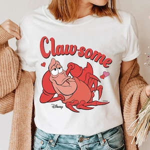 May include: White t-shirt featuring a cartoon illustration of a red crab with the text "Claw-some" in red. The design includes two pink hearts and the Disney logo. The shirt is a casual wear item.