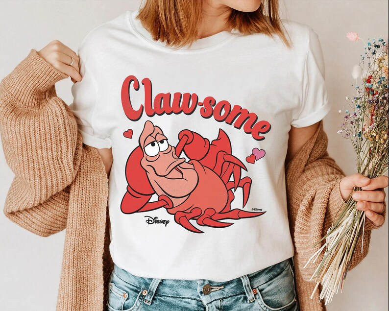 May include: White t-shirt featuring a cartoon illustration of a red crab with the text "Claw-some" in red. The design includes two pink hearts and the Disney logo. The shirt is a casual wear item.