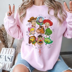 Puede incluir: Una sudadera rosa claro con un gr&aacute;fico colorido de personajes de Toy Story dispuestos como globos. Los personajes incluyen a Woody, Buzz Lightyear, Jessie y otros. Los nombres de los personajes est&aacute;n escritos en una fuente de guion.