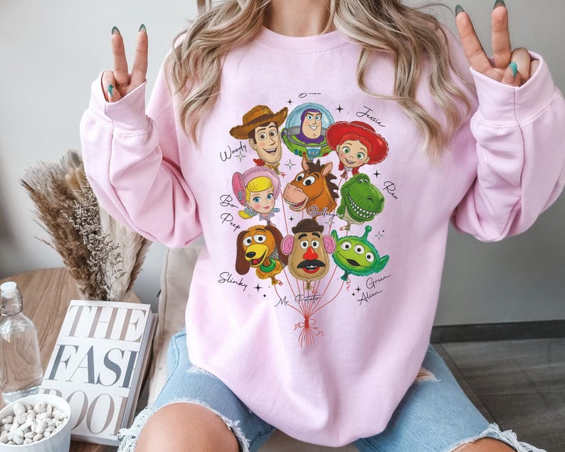 Puede incluir: Una sudadera rosa claro con un gr&aacute;fico colorido de personajes de Toy Story dispuestos como globos. Los personajes incluyen a Woody, Buzz Lightyear, Jessie y otros. Los nombres de los personajes est&aacute;n escritos en una fuente de guion.