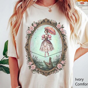 May include: Ivory Comfort Color t-shirt featuring a whimsical illustration. The design shows a woman with an umbrella, standing on a tightrope above an alligator in a pond, framed by roses. The shirt is a light cream color.