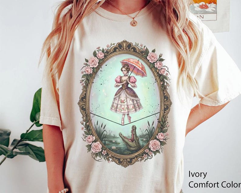 May include: Ivory Comfort Color t-shirt featuring a whimsical illustration. The design shows a woman with an umbrella, standing on a tightrope above an alligator in a pond, framed by roses. The shirt is a light cream color.