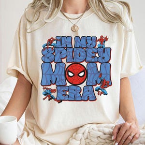 May include: Cream-colored t-shirt featuring the text "IN MY SPIDEY MOM ERA" in blue bubble letters with a red Spider-Man mask in the center. Spider-Man figures are positioned around the text.
