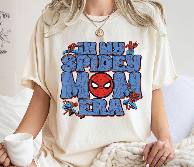 May include: Cream-colored t-shirt featuring the text "IN MY SPIDEY MOM ERA" in blue bubble letters with a red Spider-Man mask in the center. Spider-Man figures are positioned around the text.