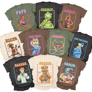 May include: A collection of t-shirts featuring various Muppet characters, including Kermit, Miss Piggy, and Fozzie Bear. Each shirt displays a character illustration within a retro-style frame, with the character's name above. The shirts are in various colors.