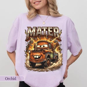 May include: Orchid-colored t-shirt featuring a cartoon image of Mater from the Cars movie. The graphic includes the word "MATER" in a metallic, distressed font above the character. The image shows Mater kicking up dust.