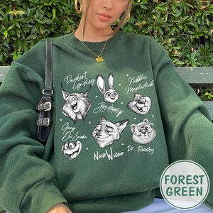Puede incluir: Sudadera verde bosque con ilustraciones de personajes de dibujos animados y texto. El dise&ntilde;o incluye personajes como Pawbert Lyndey, Judy Hopps y Nick Wilde. La sudadera tiene cuello redondo y mangas largas.