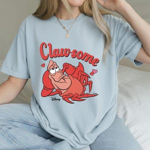 May include: Light blue t-shirt featuring a cartoon illustration of a red crab with the text "Claw-some" in red. The crab is lying down with a relaxed expression. Two small hearts are on the shirt. Disney logo is at the bottom.