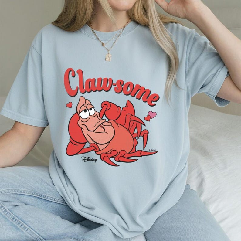 May include: Light blue t-shirt featuring a cartoon illustration of a red crab with the text "Claw-some" in red. The crab is lying down with a relaxed expression. Two small hearts are on the shirt. Disney logo is at the bottom.