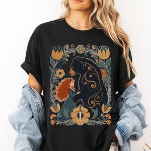 Op de afbeelding: Zwart T-shirt met een afbeelding van een beer en een vrouw met rood haar, omringd door bloemen in geel, blauw en groen. Het ontwerp bevat een zwaard en wordt omlijst door een bloemenrand.