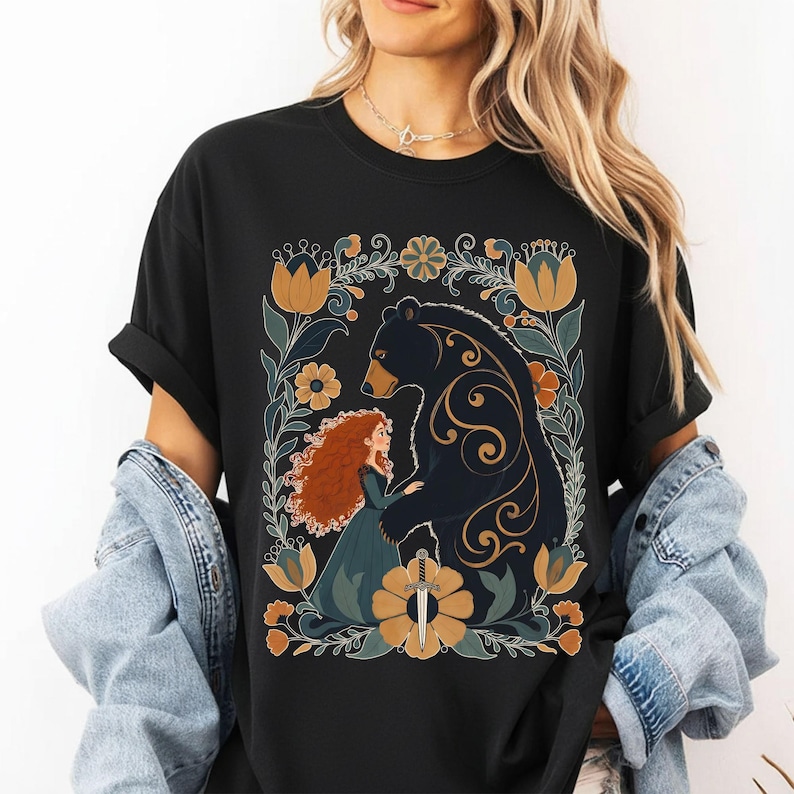 Op de afbeelding: Zwart T-shirt met een afbeelding van een beer en een vrouw met rood haar, omringd door bloemen in geel, blauw en groen. Het ontwerp bevat een zwaard en wordt omlijst door een bloemenrand.