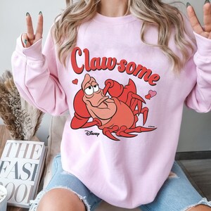 May include: A light pink sweatshirt featuring the text "Claw-some" in red script above a cartoon illustration of a red crab. The crab is from Disney's "The Little Mermaid" and is surrounded by small pink hearts.