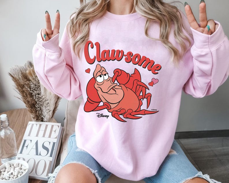 May include: A light pink sweatshirt featuring the text "Claw-some" in red script above a cartoon illustration of a red crab. The crab is from Disney's "The Little Mermaid" and is surrounded by small pink hearts.