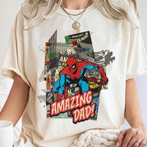 May include: Cream-colored t-shirt featuring a colorful graphic of Spider-Man in a comic book style. The design includes the words "AMAZING DAD!" in red and white, with comic book panels in the background.
