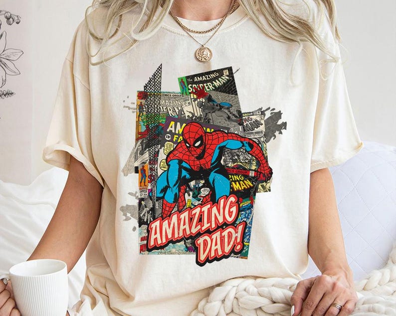 May include: Cream-colored t-shirt featuring a colorful graphic of Spider-Man in a comic book style. The design includes the words "AMAZING DAD!" in red and white, with comic book panels in the background.