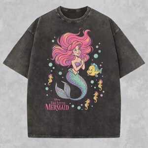 May include: A charcoal gray acid-wash t-shirt featuring a graphic of Ariel from The Little Mermaid with pink hair, a green tail, and a purple seashell top. The design includes a yellow fish, several small seahorses, bubbles, and the text The Little Mermaid.