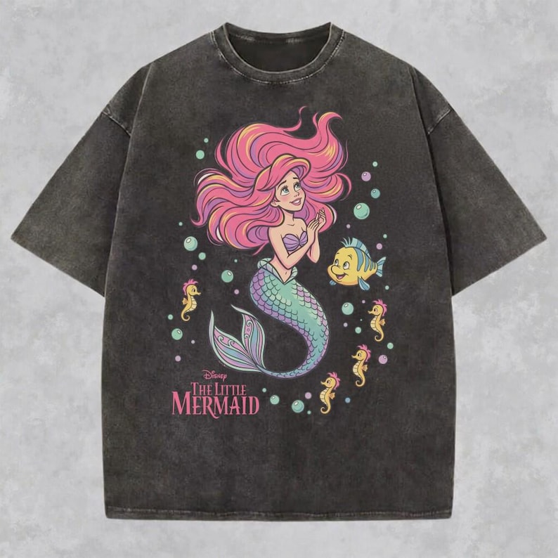 May include: A charcoal gray acid-wash t-shirt featuring a graphic of Ariel from The Little Mermaid with pink hair, a green tail, and a purple seashell top. The design includes a yellow fish, several small seahorses, bubbles, and the text The Little Mermaid.