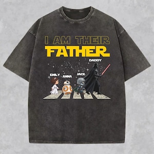 May include: A charcoal grey short-sleeve t-shirt featuring a graphic print with the text I AM THEIR FATHER and DADDY. The design shows cartoon characters walking on a zebra crossing against a starry background, including names Emily, Anna, and Jack.