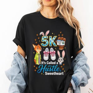May include: Black t-shirt with a colourful graphic featuring cartoon characters, running shoes, and the text "5K It's Called a Hustle, Sweetheart." The design includes paw prints and the words "Springtime Surprise 2026."