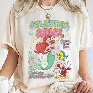 May include: A cream-colored t-shirt featuring a graphic print of Ariel the mermaid, Flounder, and Sebastian. The text reads Atlantica Ariel Ocean Tour 1991 and Sebastian with the Hot Crustacean Band. The design includes colorful bubbles, coral, and musical notes.