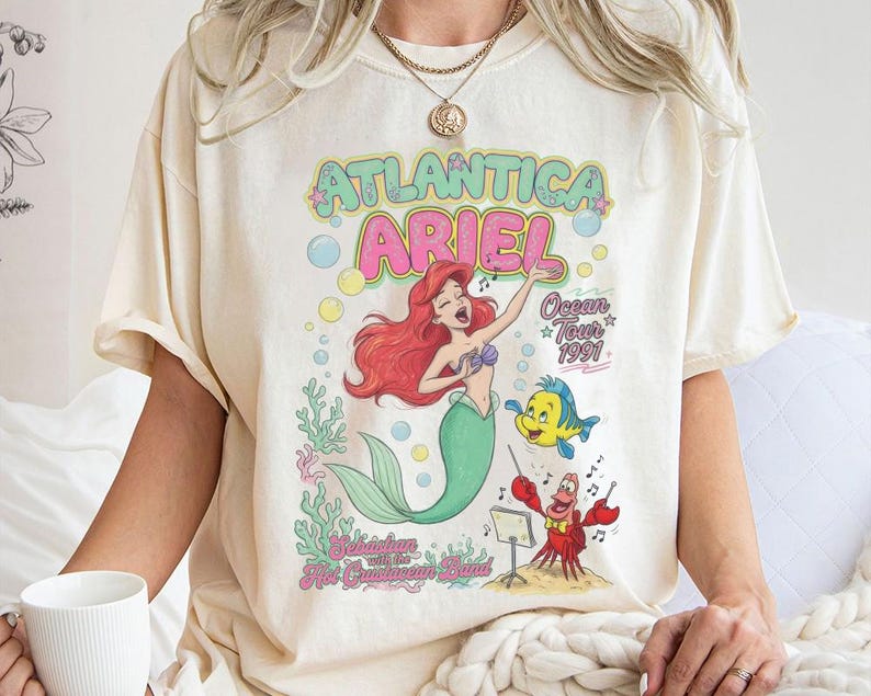 May include: A cream-colored t-shirt featuring a graphic print of Ariel the mermaid, Flounder, and Sebastian. The text reads Atlantica Ariel Ocean Tour 1991 and Sebastian with the Hot Crustacean Band. The design includes colorful bubbles, coral, and musical notes.