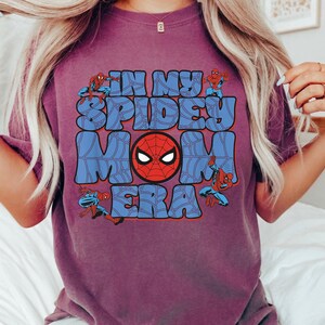 May include: A purple t-shirt with the text "IN MY SPIDEY MOM ERA" in blue bubble letters. The design includes a red Spider-Man mask and Spider-Man figures in action poses.