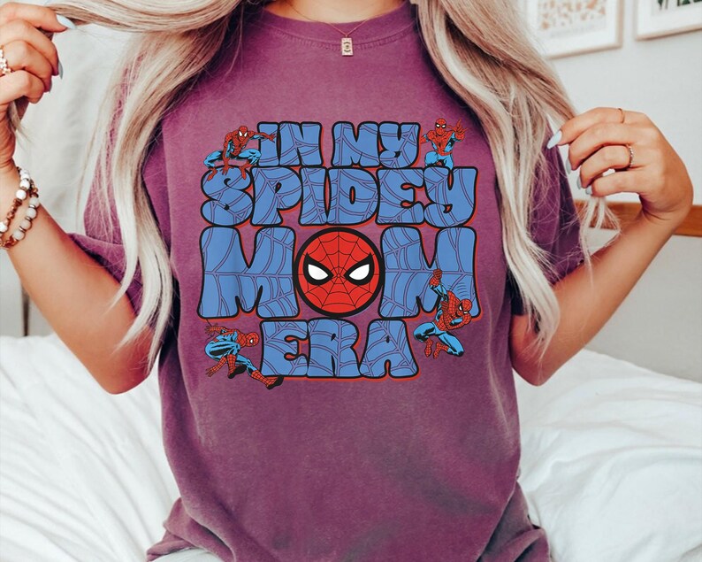 May include: A purple t-shirt with the text "IN MY SPIDEY MOM ERA" in blue bubble letters. The design includes a red Spider-Man mask and Spider-Man figures in action poses.