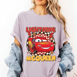 May include: A light purple t-shirt featuring a graphic of Lightning McQueen from the movie Cars. The design includes the text "LIGHTNING MCQUEEN" in red and yellow, with a checkered background and lightning bolt accents.
