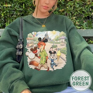 May include: Forest green sweatshirt with a graphic of cartoon characters Nick Wilde and Judy Hopps from Zootopia, holding treats in a park setting. The design includes a castle, food carts, and balloons. The sweatshirt has a crew neck.