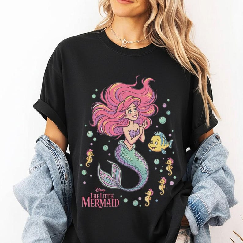 May include: A black short-sleeved t-shirt featuring a graphic print of Ariel from The Little Mermaid with pink hair, a green tail, and a yellow fish companion. The design includes small seahorses, bubbles, and the text The Little Mermaid.