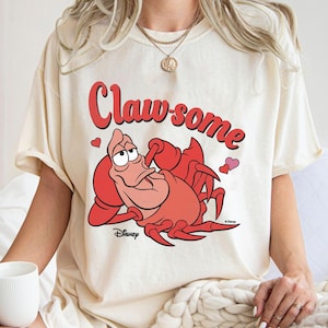 May include: Cream-colored t-shirt featuring a cartoon illustration of Sebastian the crab from Disney's The Little Mermaid. The text "Claw-some" is written in red above the crab. Two small hearts are also included.